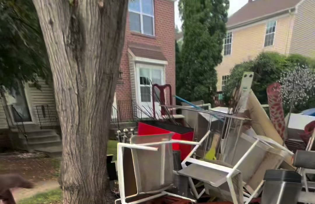 Chairs, a cabinet and a pile of household furniture stacked at the curb under a tree outside a row of brick townhouses.