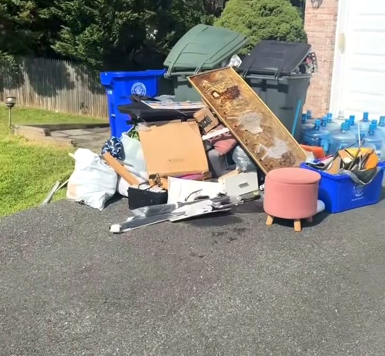 A pile of junk left at the end of a driveway: a headboard, a footstool, bagged rubbish and flattened boxes beside the trash cans.